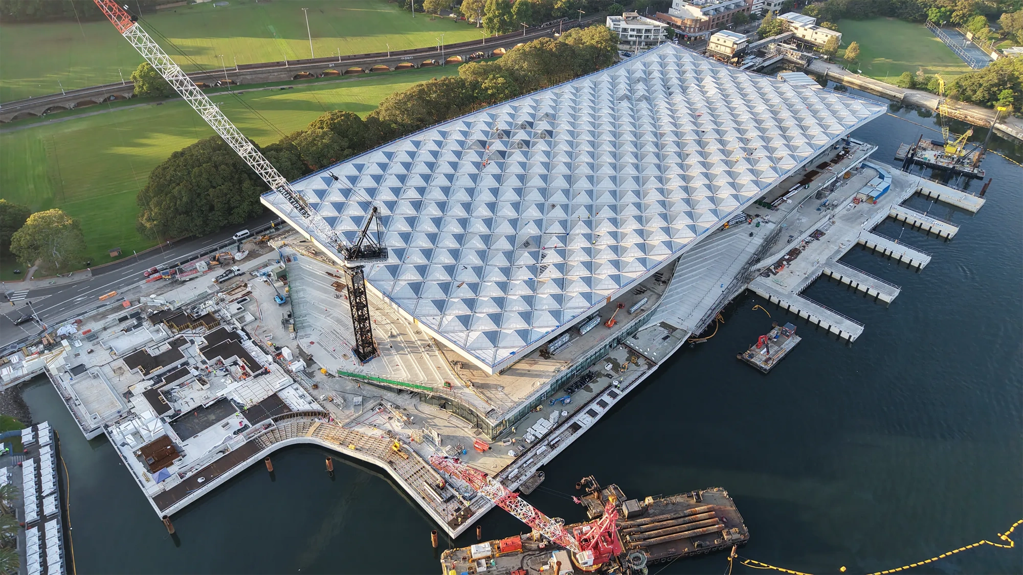 Ariel view of a large waterfront construction site with a geometric roof structure and surrounding cranes and platforms