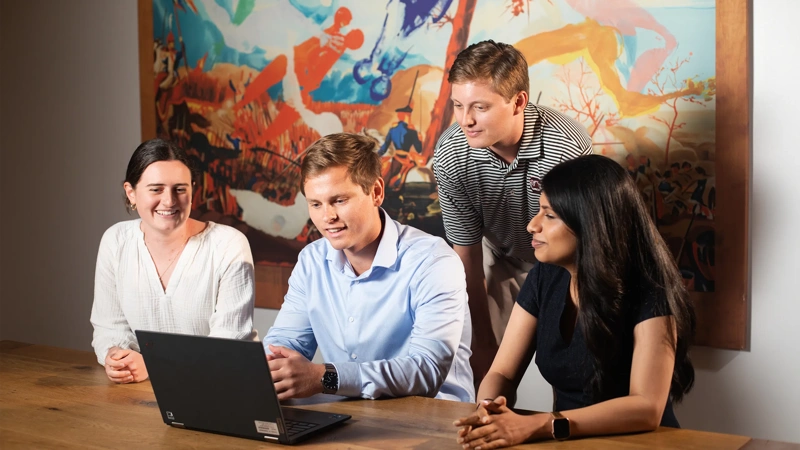 Four colleagues sitting in an office space gathered around male colleague staring at his laptop on a brown table, engaged in a lively conversation.