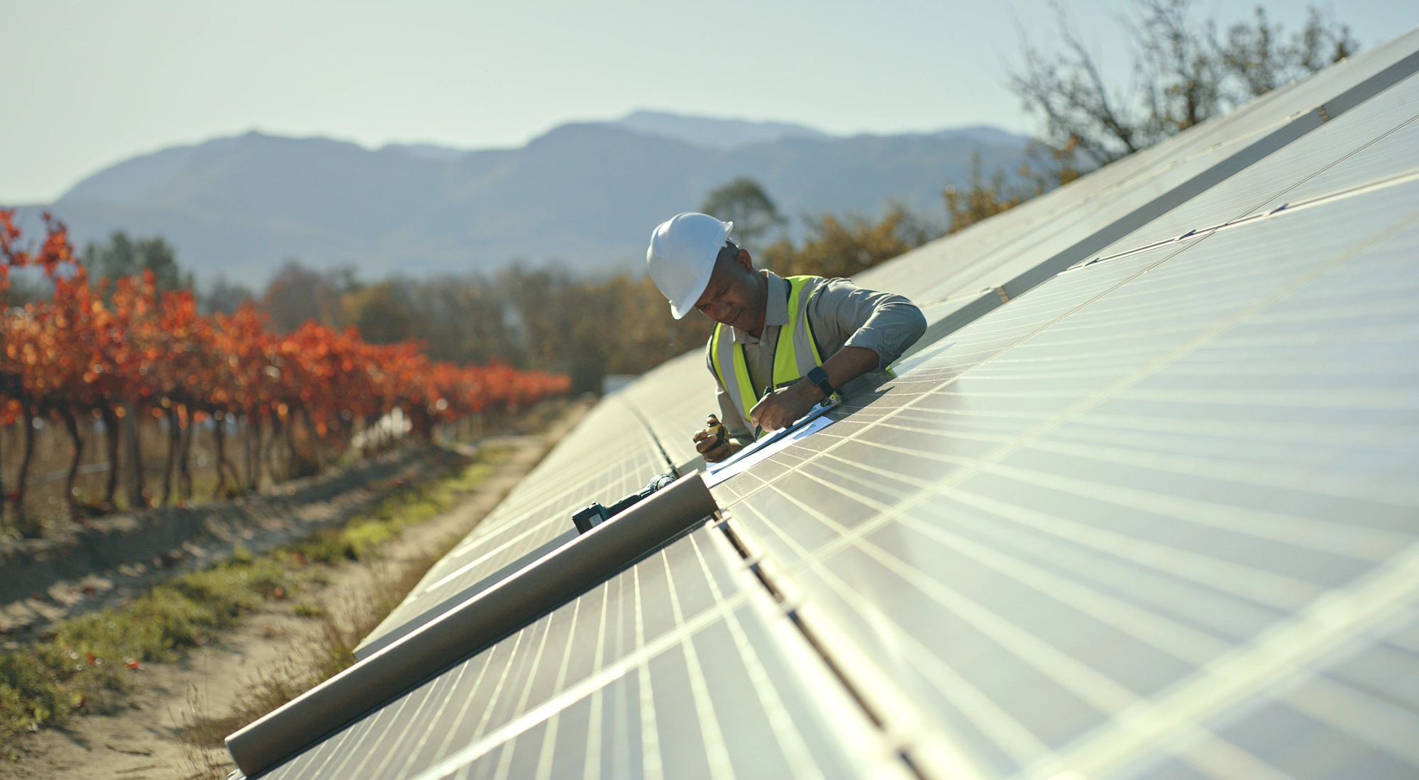 A workman working on a solar panel installation with a backdrop of a mountain and trees with orange and red leaves.