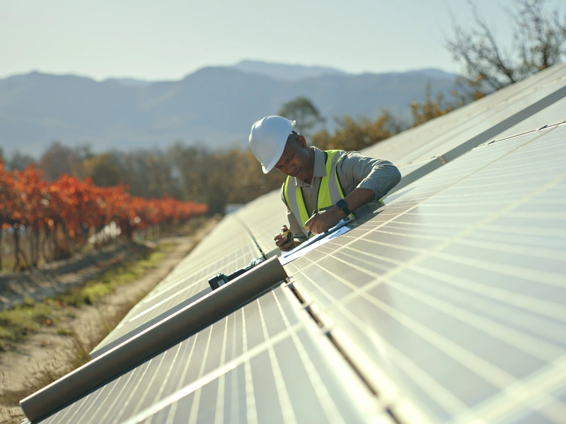 A workman working on a solar panel installation with a backdrop of a mountain and trees with orange and red leaves.
