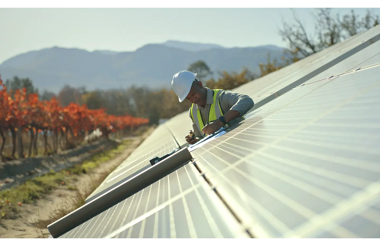 A workman working on a solar panel installation with a backdrop of a mountain and trees with orange and red leaves.  