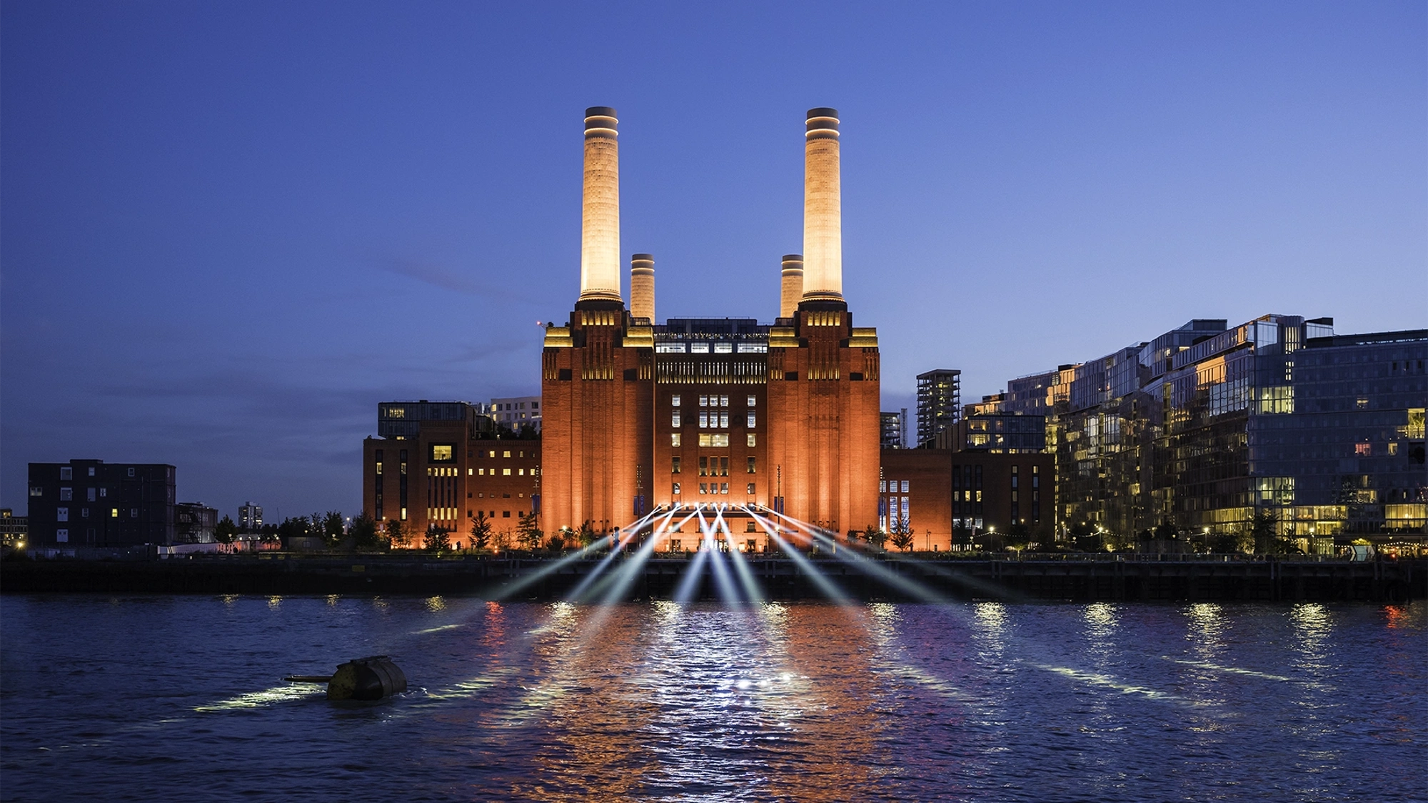 A striking image of London's iconic Battersea Power Station against a blue night sky. The industrial columns are lit up and light also shines on the water in front. To the right of the building are a block of modern, glass fronted flats.