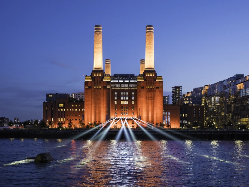 A striking image of London's iconic Battersea Power Station against a blue night sky. The industrial columns are lit up and light also shines on the water in front. To the right of the building are a block of modern, glass fronted flats.