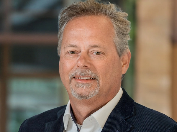 Portrait of Ron Staa, Director, outside in front of a blurred brown background featuring a building and mix of natural and urban elements.