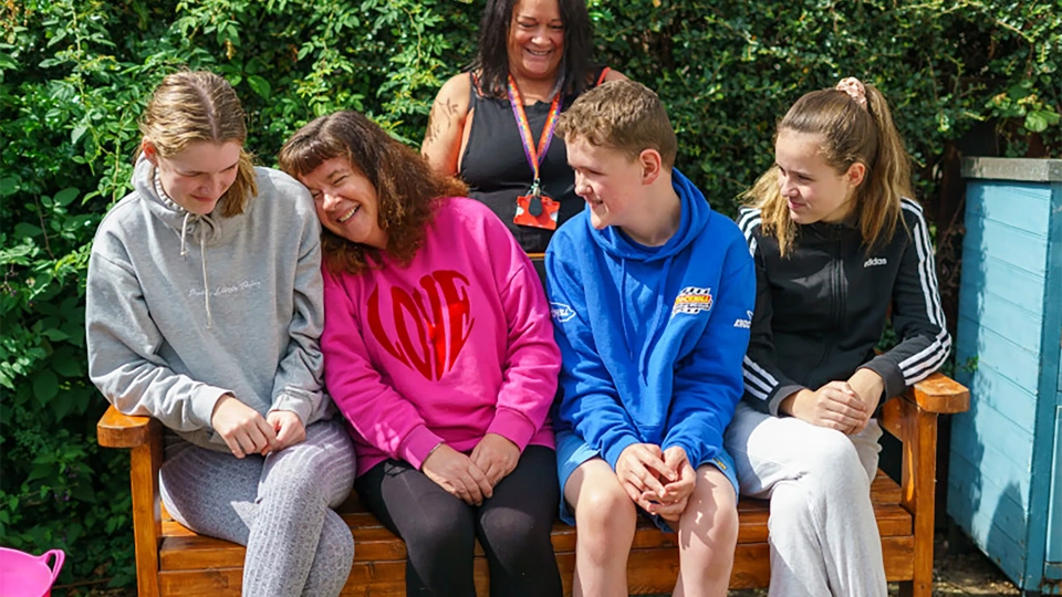 Picture of four children sitting on a bench outside in pink, blue, black and grey clothing, with a woman standing behind the bench.