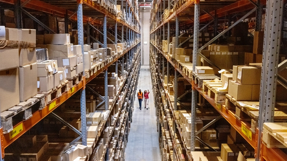 Two employees in high vis jackets walking through warehouse with tall shelves full of products