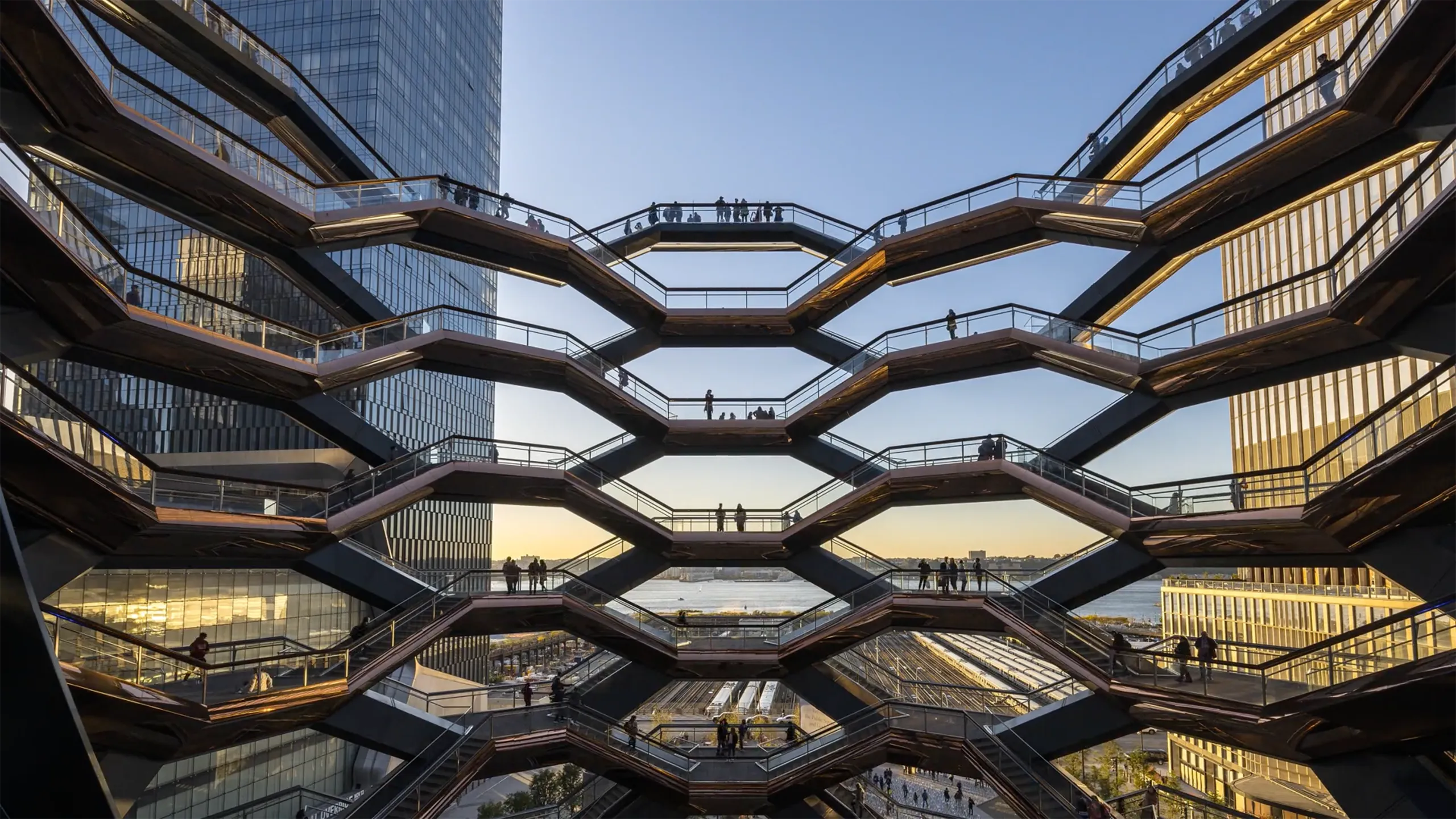 Architectural view of the Vessel at Hudson Yards in New York City, showing multiple levels of interconnected staircases with silhouetted people walking against a backdrop of modern skyscrapers and a sunset sky