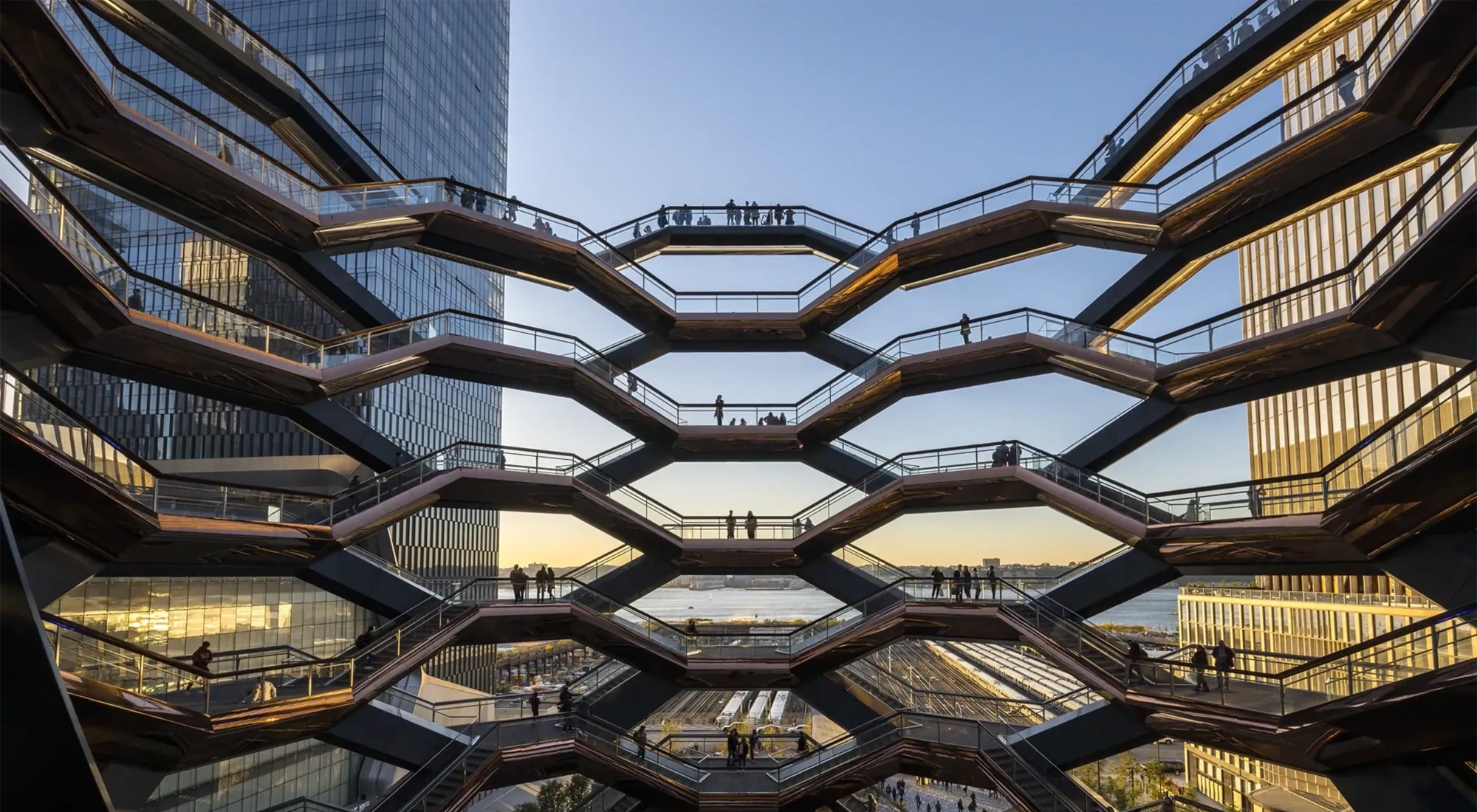Architectural view of the Vessel at Hudson Yards in New York City, showing multiple levels of interconnected staircases with silhouetted people walking against a backdrop of modern skyscrapers and a sunset sky