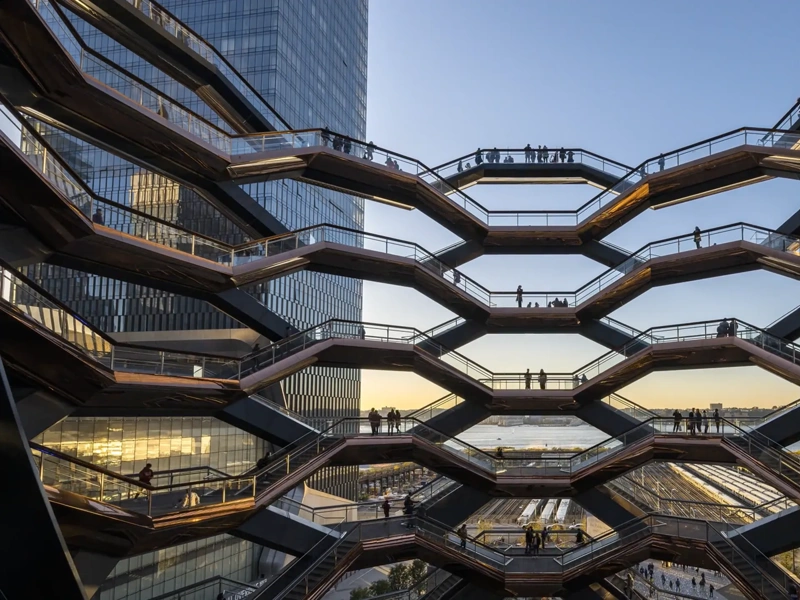 Architectural view of the Vessel at Hudson Yards in New York City, showing multiple levels of interconnected staircases with silhouetted people walking against a backdrop of modern skyscrapers and a sunset sky
