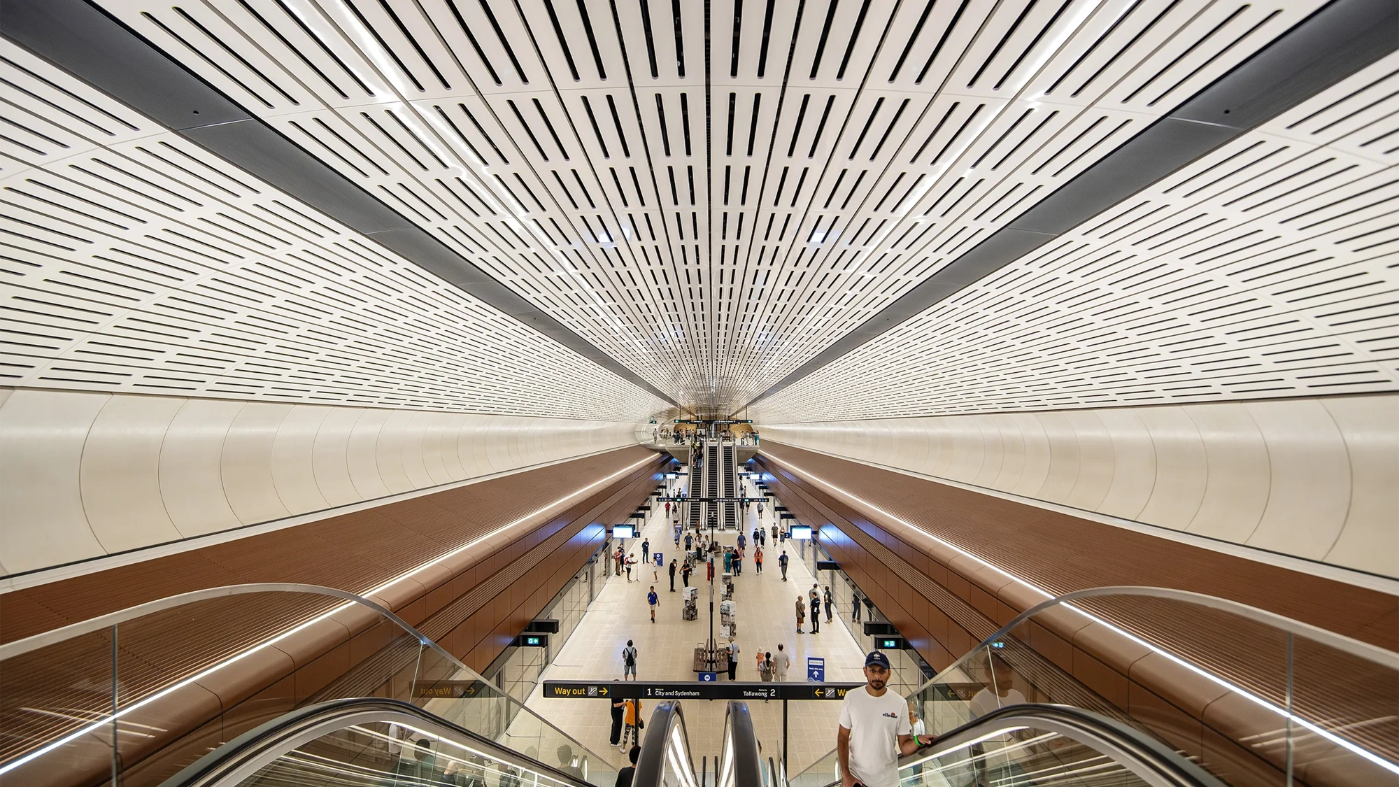 Interior view of Sydney metro modern underground station with a wide ceiling featuring linear ventilation panels, symmetrical architecture and passengers moving through space below.