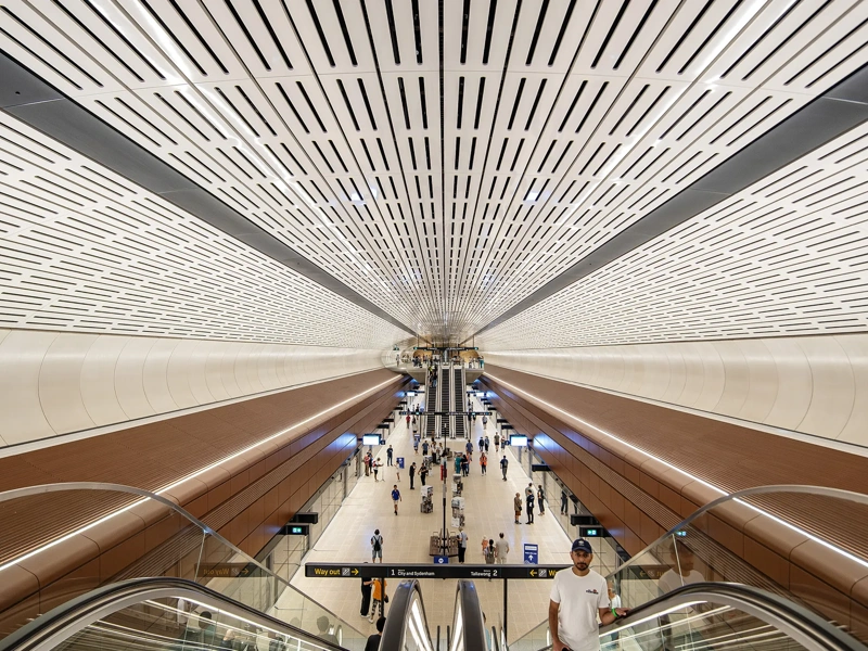 Interior view of Sydney metro modern underground station with a wide ceiling featuring linear ventilation panels, symmetrical architecture and passengers moving through space below.