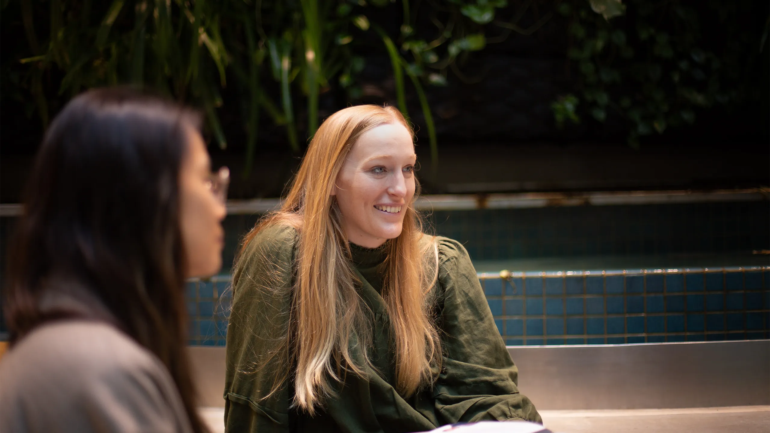 Two colleagues together with the main focus featuring a blonde woman having a conversation in a plant-filled office