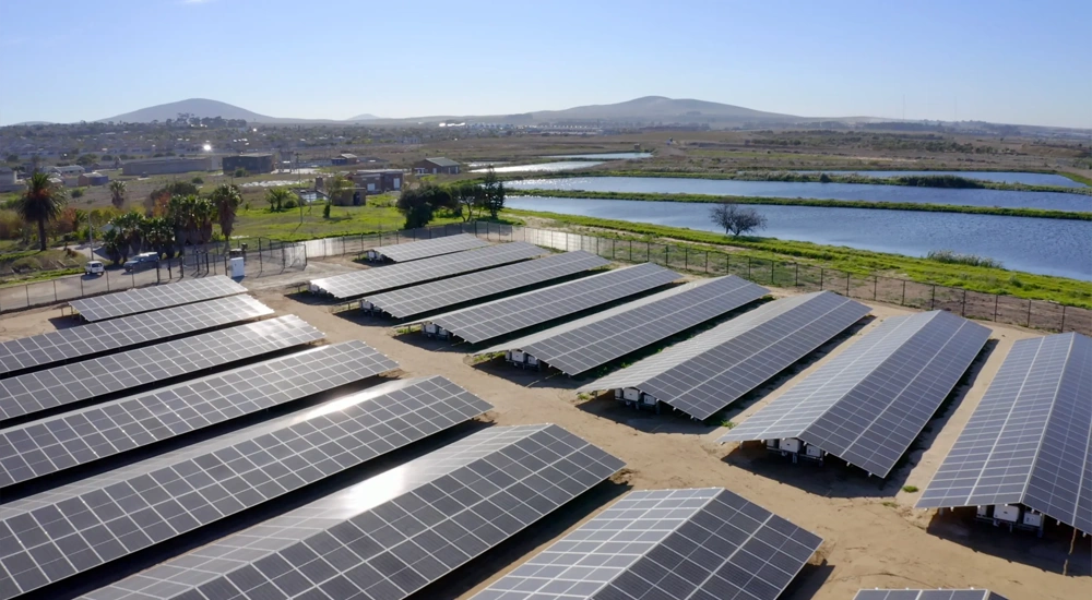 A collection of solar panels next to a body of water with mountains in the background.