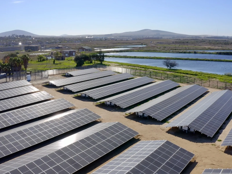 A collection of solar panels next to a body of water with mountains in the background.