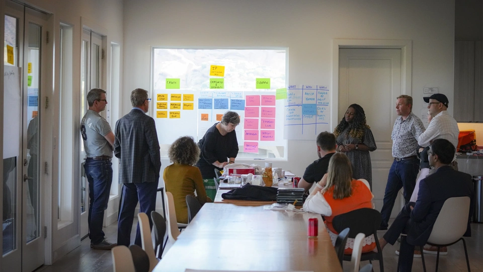 Photo of a group of people seated and standing around a table in a boardroom during a meeting, with a project screen behind them displaying different coloured blocks of text in the form of an organisational chart