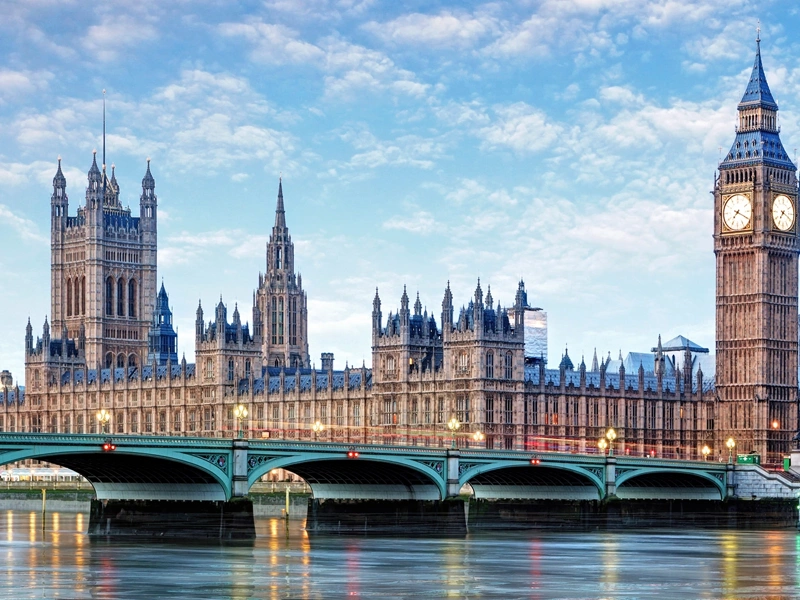 Houses of Parliament, in London, England. Taken from across the River Thames, capturing the entirety of the historic building, including the Elizabeth Tower, which houses Big Ben, on the right. The Westminster Bridge spans the river in the foreground, with the calm water reflecting the structure above. The sky is partly cloudy, and the scene is bathed in soft daylight.