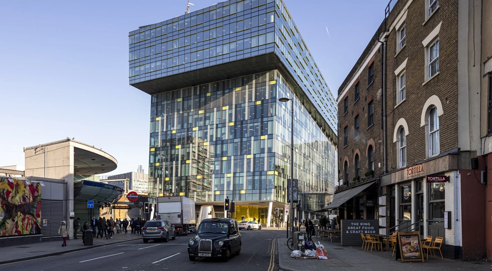 Modern urban street scene in London, featuring a striking contemporary glass building with a cantilevered upper section. This building is set against a backdrop of older brick structures, including a corner restaurant named "Tortilla" with outdoor seating. A classic black London taxi and other vehicles are visible on the street, along with pedestrians walking by. The area appears busy with a mix of architectural styles, reflecting both modern and historical elements of the city.