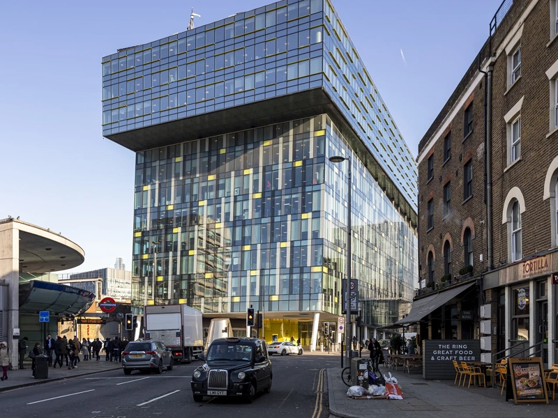 Modern urban street scene in London, featuring a striking contemporary glass building with a cantilevered upper section. This building is set against a backdrop of older brick structures, including a corner restaurant named "Tortilla" with outdoor seating. A classic black London taxi and other vehicles are visible on the street, along with pedestrians walking by. The area appears busy with a mix of architectural styles, reflecting both modern and historical elements of the city.