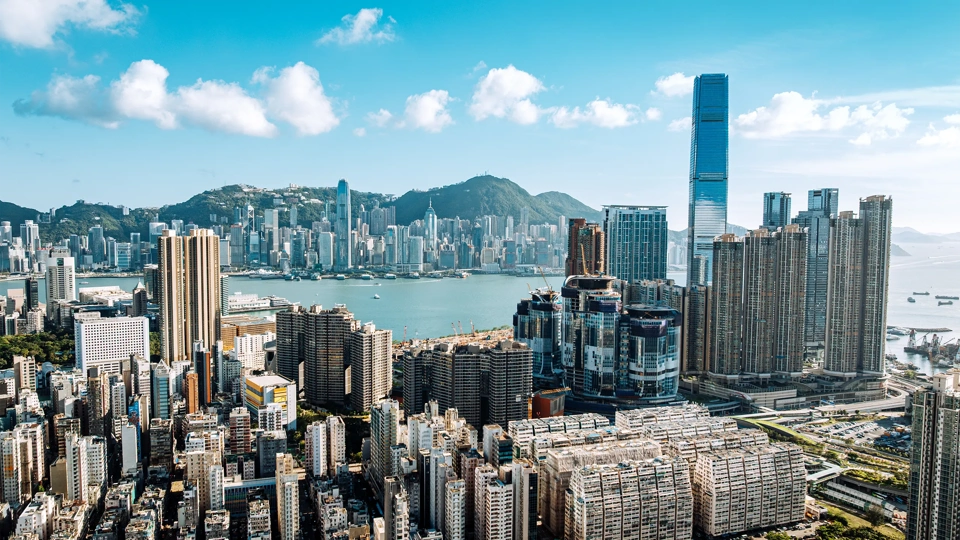 Urban skyline of Hong Kong city featuring high-rise buildings and the harbor surrounded by mountains and blue skies.