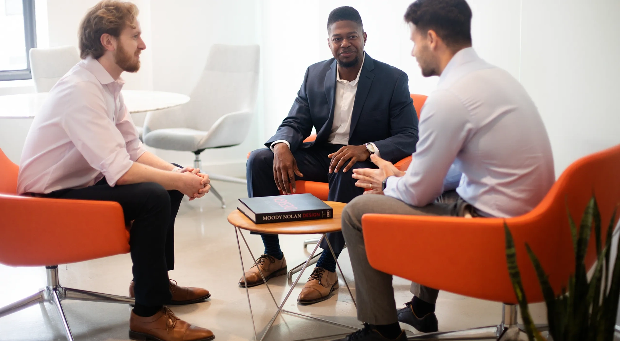 Three male colleagues sitting down on bright orange chairs, engaging in conversation. The office environment is bright and modern with plants behind the chairs.