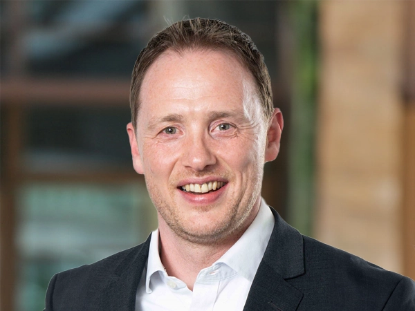 Portrait of Garry Murphy, UK Head of Project Management, Infrastructure, outside in front of a blurred brown background featuring a building and mix of natural and urban elements.