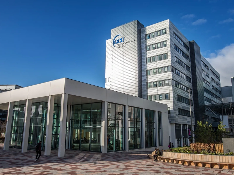 Entrance of Glasgow Caledonian University. Silver building with glass windows and brown bench outside against blue sky.