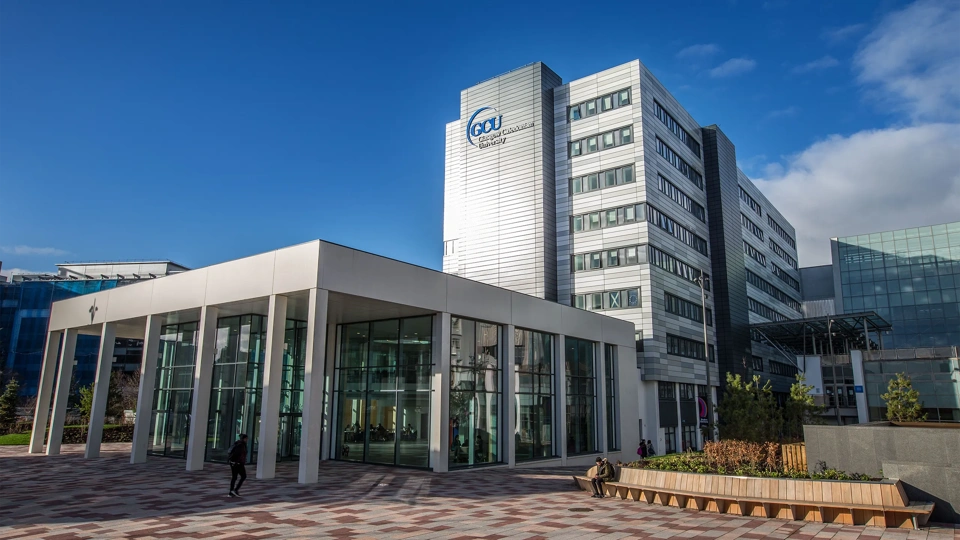 Entrance of Glasgow Caledonian University. Silver building with glass windows and brown bench outside against blue sky.