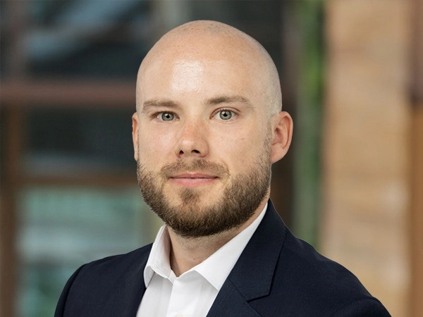 Portrait of Kevin Higgins, Lead Cost Manager, outside in front of a blurred brown background featuring a building and mix of natural and urban elements.