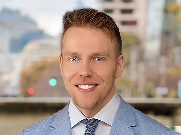 Portrait of Nicholas Rita, Associate Director, standing outside featuring an urban cityscape with tall buildings and a mix of architectural styles. The scene includes some trees with autumn foliage, adding a touch of color. 