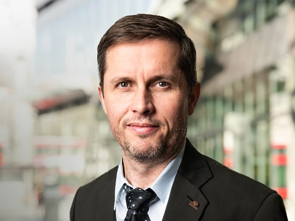 Morten Søjborg standing outside in front of buildings featuring an urban cityscape with a mix of architectural styles. The scene includes some windows and red features, adding a touch of color.