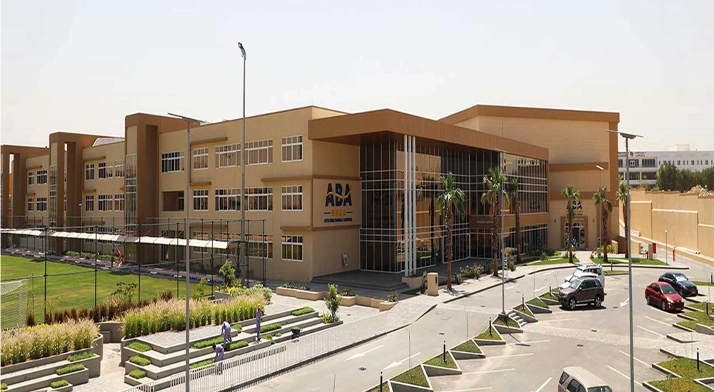 A sand coloured block of school buildings with a car park in front and small playing field to the left of the building.