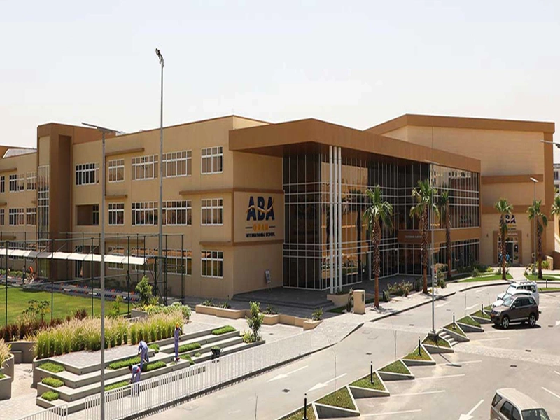 A sand coloured block of school buildings with a car park in front and small playing field to the left of the building.