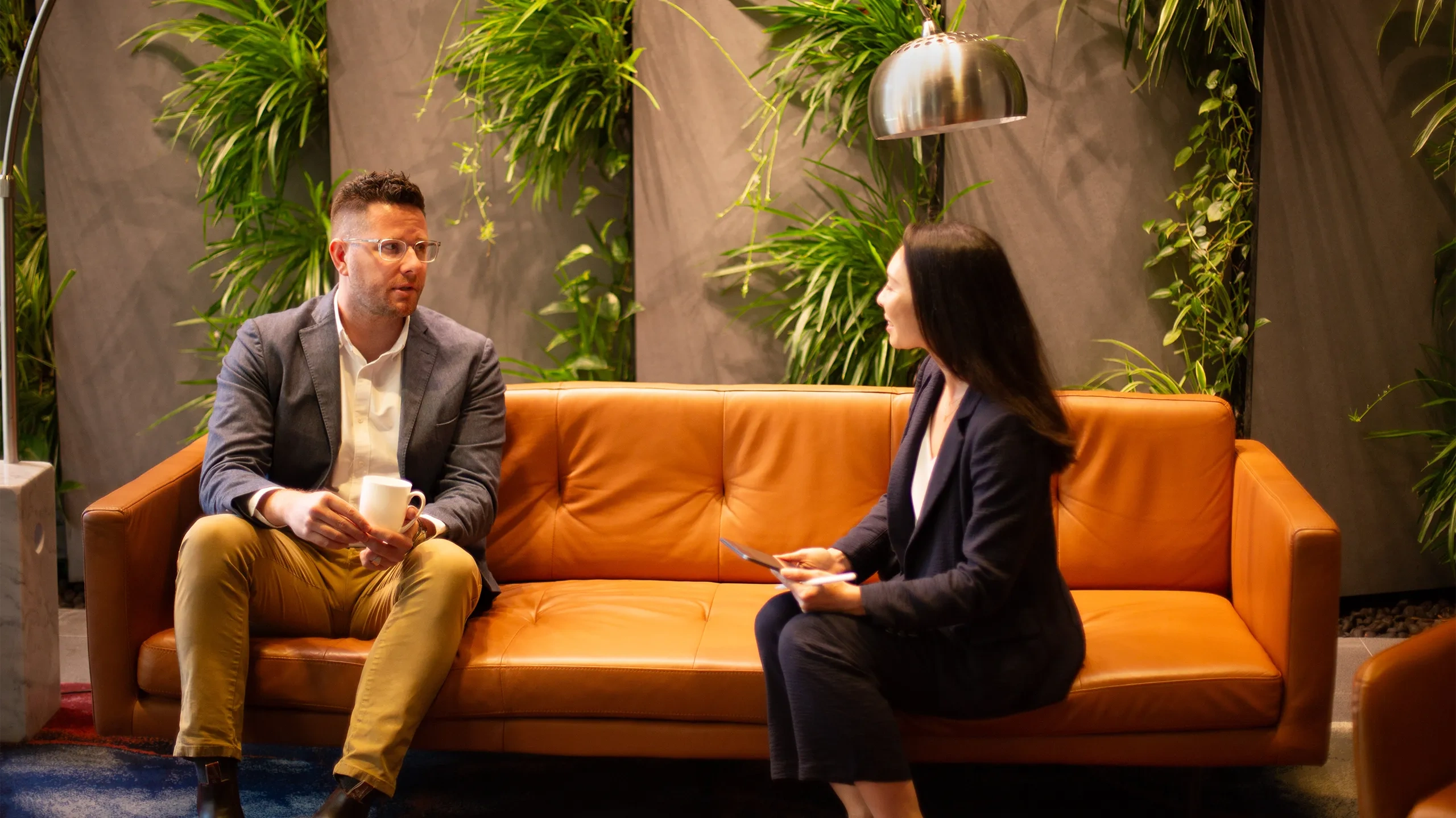 Male and female colleagues sitting together on orange sofas in a plant-filled office having a discussion.