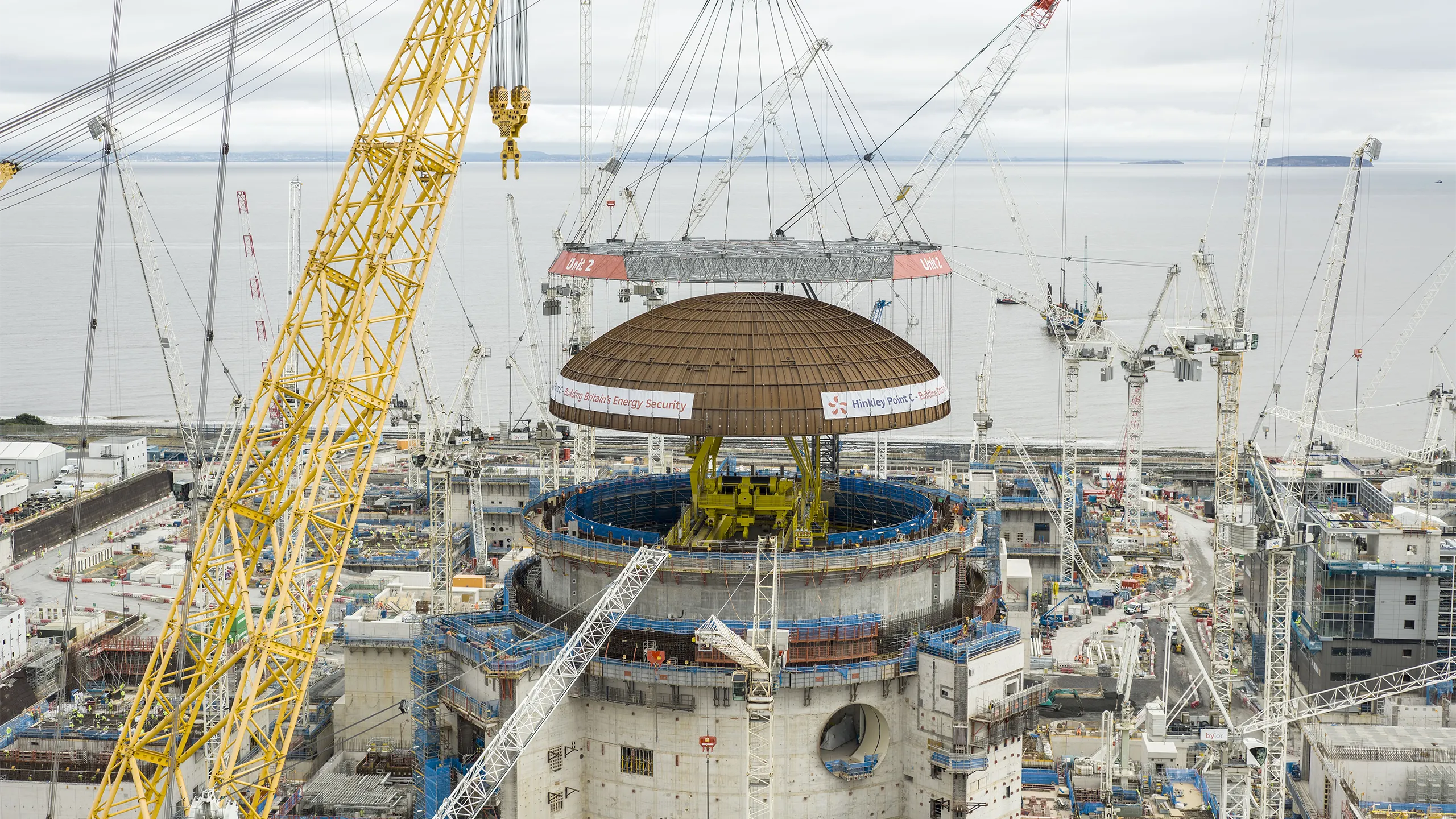 A large crane lifts a domed steel structure into place above a major construction site with multiple cranes and industrial buildings.