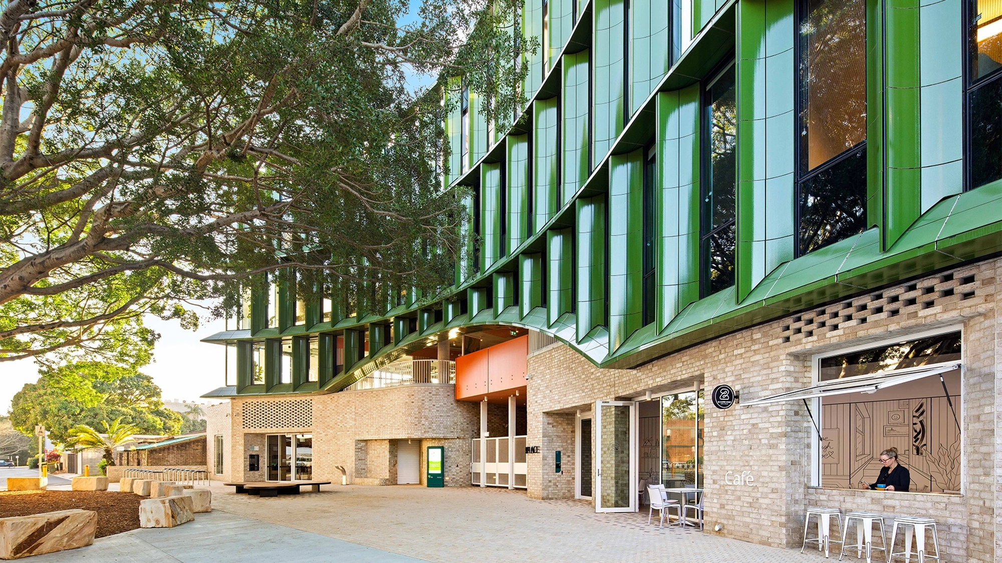The exterior of a modern, green building with foliage and seating next to it.
