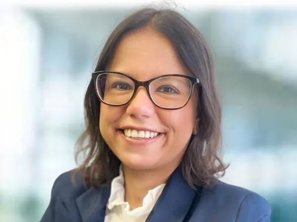 Portrait of Monica Pontes, Communications Manager, smiling in blue suit against blurry blue / grey office background.