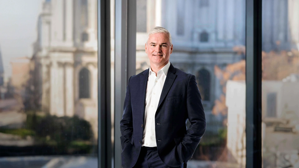 Vincent Clancy standing in the London office with the London city view in the background