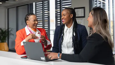 Three female colleagues in office space gathered around laptop, smiling and engaging in conversation.