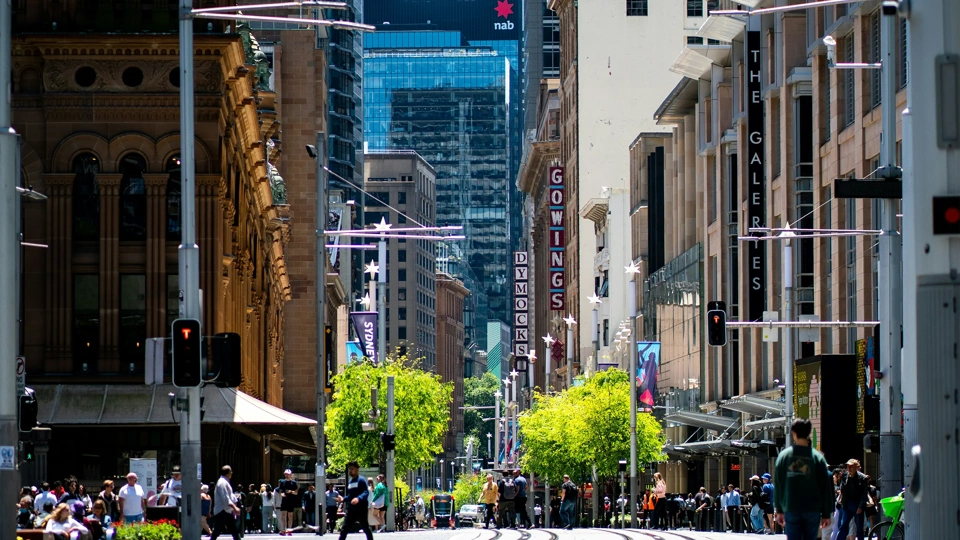 City landscape with people walking and trees in the center with tram coming down in distance.