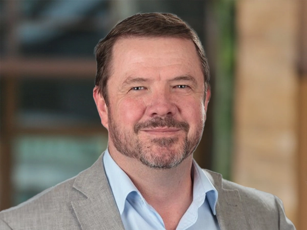 Portrait of Liam Hale, Defence Sector Lead, outside in front of a blurred brown background featuring a building and mix of natural and urban elements.
