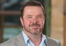 Portrait of Liam Hale, Defence Sector Lead, outside in front of a blurred brown background featuring a building and mix of natural and urban elements.