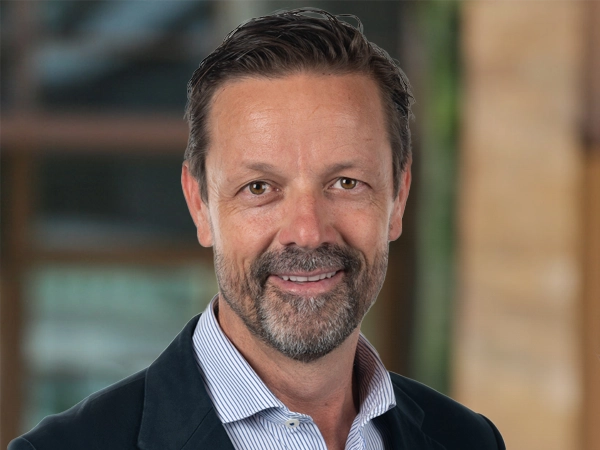 Mark McDonald, Project Director, Infrastructure, outside in front of a blurred brown background featuring a building and mix of natural and urban elements.
