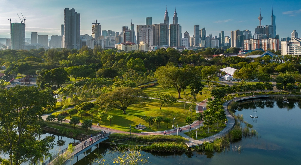 Aerial view of a city park with lush greenery, walking paths and a lake, set against a backdrop of tall sky scrappers