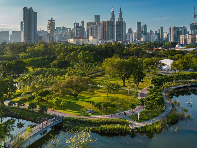 Aerial view of a city park with lush greenery, walking paths and a lake, set against a backdrop of tall sky scrappers