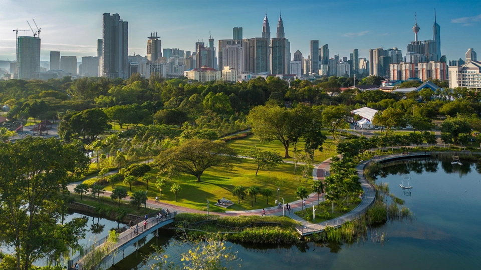 Aerial view of a city park with lush greenery, walking paths and a lake, set against a backdrop of tall sky scrappers