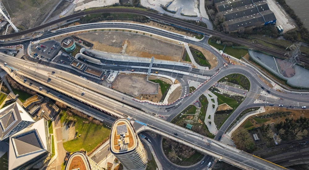 View of constructed city overpass alongside grass and trees with modern high rise buildings in the background.